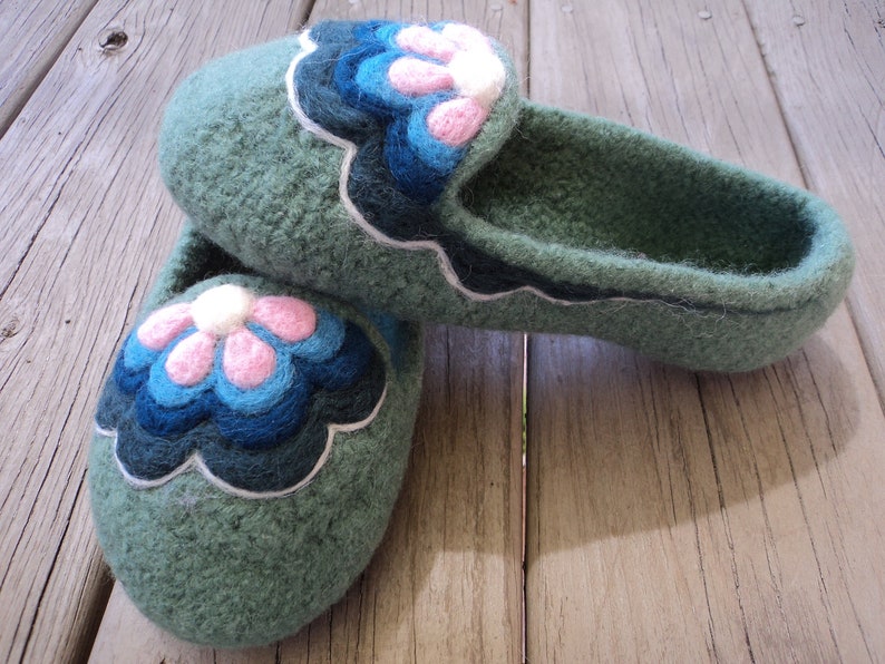 Puede incluir: Un par de zapatillas de lana afieltrada en color verde salvia con una aplicaci&oacute;n floral festoneada en la parte superior. El dise&ntilde;o de la flor consta de p&eacute;talos superpuestos en tonos rosa claro, azul brillante y azul marino, con un borde blanco.