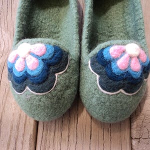 Puede incluir: Un par de zapatillas de lana afieltrada en color verde salvia con una aplicaci&oacute;n floral decorativa en la puntera. La flor consta de p&eacute;talos superpuestos en tonos rosa, azul claro y azul oscuro, delineados con un borde blanco sobre la base verde.