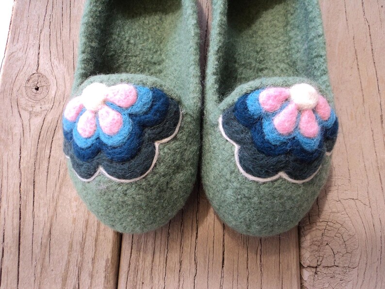 Puede incluir: Un par de zapatillas de lana afieltrada en color verde salvia con una aplicaci&oacute;n floral decorativa en la puntera. La flor consta de p&eacute;talos superpuestos en tonos rosa, azul claro y azul oscuro, delineados con un borde blanco sobre la base verde.