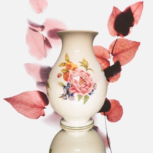 May include: A white ceramic vase with a floral design featuring pink, yellow, and green flowers. The vase has a round body and a flared top. It is sitting on a white pedestal.