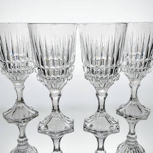 May include: Four clear crystal wine glasses with detailed vertical and geometric cut patterns. The glasses feature a classic design with a wide bowl, a stem, and a decorative base. The glasses are displayed on a reflective surface.