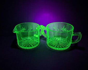 May include: Two vintage green glass cream and sugar set. The pieces have handles and are decorated with etched leaf and starburst patterns. The glass glows under a black light, showcasing its unique color and design. The set is displayed against a dark background.