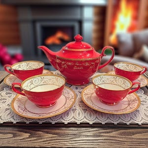 May include: A bright red tea set consisting of a teapot with a lid and four matching teacups on saucers. Each piece features a white interior and a delicate gold filigree scroll pattern along the rims and sides, set on a white lace mat.