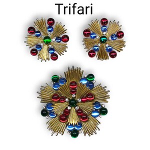 May include: A set of three gold-toned Trifari brooches featuring a starburst design with red, blue, and green glass stones.