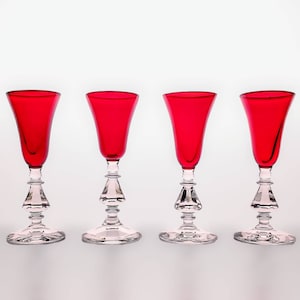 May include: Four red glass goblets with clear glass stems and bases. The goblets are arranged in a row on a white surface.