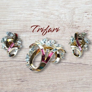 Op de afbeelding: Een vintage Trifari sieradenset met een broche en bijpassende clip-on oorbellen. De set heeft een goudkleurige metalen basis met heldere en roze strass-steentjes. De broche en oorbellen hebben een bloemmotief.