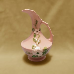 May include: A pink ceramic pitcher with a floral design. The pitcher has a wide mouth and a handle. The flowers are white with green leaves.