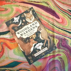 May include: A dark green oracle deck titled "Woodland Wardens" featuring illustrations of foxes and flowers. The deck includes a 52-card oracle deck and guidebook. The author's name, Jessica Roux, is printed at the bottom.