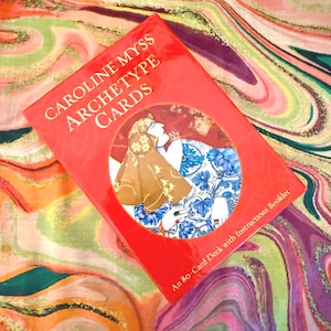 May include: A red box of "Caroline Myss Archetype Cards" with an illustration of two figures in a circle. The text "An 80-Card Deck with Instructions Booklet" is at the bottom. The background is a colorful, swirled fabric.