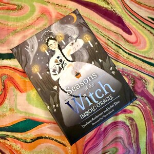 May include: A book titled "Seasons of the Witch: Imbolc Oracle" with an illustration of a woman in a white dress and long braid, surrounded by candles and a crescent moon. The cover is dark with white text.