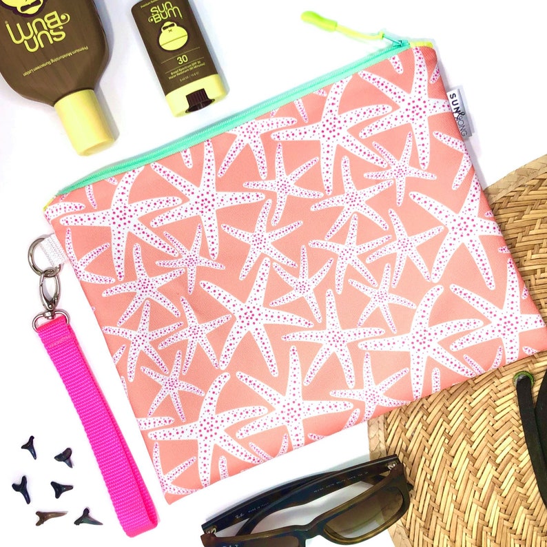 May include: A pink zippered pouch with a white starfish pattern and a pink strap. The pouch is lying on a white surface with a straw hat, sunglasses, and two bottles of sunscreen.