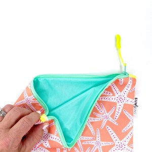 May include: A coral-coloured zippered pouch with a turquoise lining. The pouch is decorated with a white starfish pattern and has a bright yellow zipper pull. The pouch has the text "SUN & SONG" printed on the side.