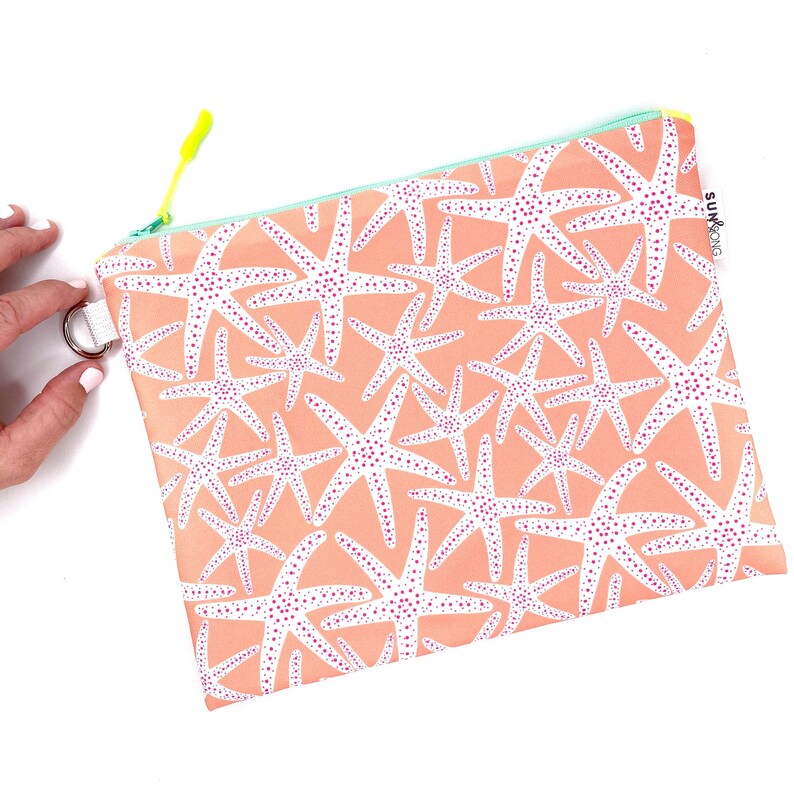 May include: A peach-coloured zippered pouch with a white starfish pattern and pink polka dots. The pouch has a neon green zipper pull and a white zipper. The pouch has a white tag with the text "SUN & KONG" on it.