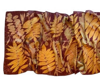 May include: A maroon silk scarf with a botanical print of yellow and brown leaves. The scarf is folded and draped on a white background.