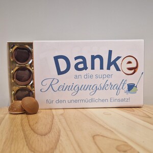 May include: A box of chocolates with a white lid featuring blue text that reads "Danke" and "Reinigungskraft". The box is open, revealing several chocolates. Two chocolates sit in front of the box on a wooden surface.