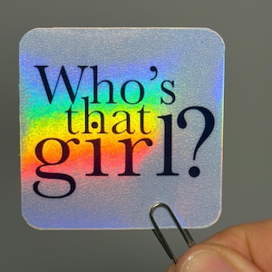 Può includere: Adesivo olografico quadrato con angoli arrotondati. L'adesivo presenta il testo "Who's that girl?" in nero e arcobaleno. Una graffetta argentata tiene l'adesivo.