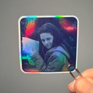 May include: Holographic sticker featuring a black and white image of a person with long hair, wearing a hooded jacket. The sticker has a square shape with rounded corners and a rainbow iridescent border. A hand holds the sticker with a silver paper clip.