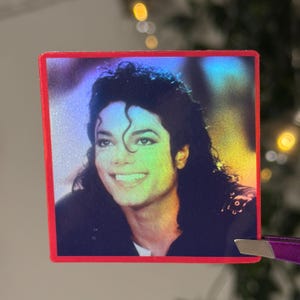 Puede incluir: Una pegatina cuadrada con borde rojo presenta un retrato sonriente de Michael Jackson. La imagen tiene un efecto holográfico, con colores cambiantes. El fondo está borroso, con toques de verde y amarillo.