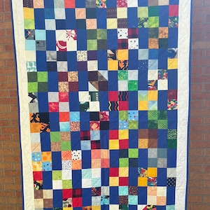 May include: A handmade patchwork quilt with a blue background and a grid of colorful squares. The squares feature various patterns and fabrics, including floral, geometric, and solid colors. The quilt is bordered with a white fabric and a blue binding.