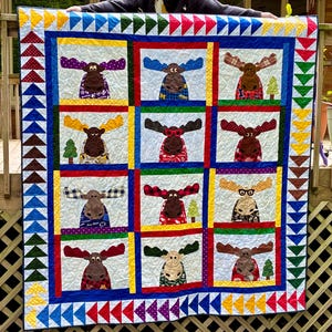 May include: A handmade quilt with a repeating pattern of moose appliqués. Each moose has unique patterned antlers and clothing. The quilt has a blue border with colourful triangle accents and measures approximately 152 cm by 152 cm.