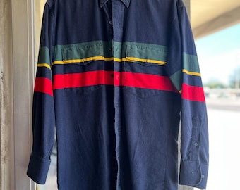 Könnte beinhalten: Ein marineblaues Langarmhemd mit Button-Down-Kragen und zwei Brusttaschen. Das Hemd hat horizontale Streifen in Grün, Gelb und Rot über Brust und Ärmeln. Das Hemd hängt an einem weißen Kleiderbügel.
