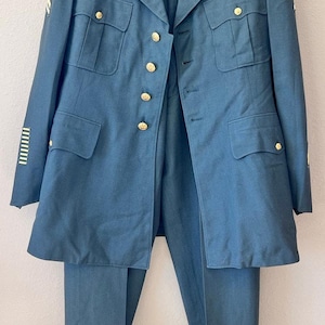 Op de afbeelding: Een vintage militair uniform in een gedempte teal kleur, bestaande uit een jas en een broek. De jas heeft goudkleurige knopen, meerdere zakken en schouderstukken. De broek heeft rechte pijpen. Het uniform wordt op een hanger getoond.