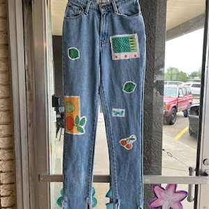 Könnte beinhalten: Hellblaue Jeans mit verschiedenen bunten Aufnähern, darunter Früchte, eine Schleife und ein Fernseher im Retro-Stil. Die Jeans haben eine hohe Taille und einen ausgefransten Saum an den Knöcheln. Die Aufnäher sind in Grün-, Orange- und Weißtönen gehalten.