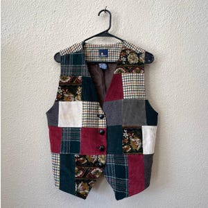 Puede incluir: Un chaleco sin mangas con un diseño de patchwork de paneles de pana, cuadros y tela floral en tonos burdeos, verde bosque, crema y gris. El chaleco tiene un cierre frontal con cuatro botones negros y un forro interior de color marrón oscuro.