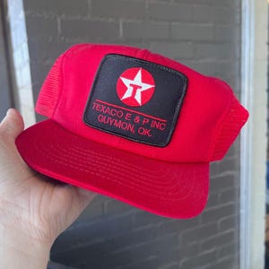 Può includere: Cappellino da camionista rosso con retro in rete e visiera curva. La parte anteriore presenta una toppa quadrata nera con il logo Texaco e la scritta "TEXACO E & P INC GUYMON, OK."