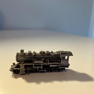 Puede incluir: Una maqueta detallada de una locomotora de tren en tonos grises y negros. El tren en miniatura presenta detalles intrincados, incluyendo una cabina, chimenea y varias tuberías y ruedas. El modelo está sobre un fondo neutro.