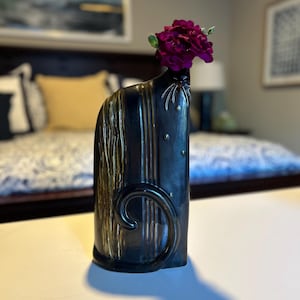 Puede incluir: Un jarrón decorativo negro con detalles dorados y un diseño en espiral. El jarrón contiene un racimo de flores de color púrpura intenso. El jarrón está sobre una superficie blanca.