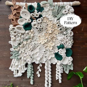 May include: A handmade macrame wall hanging with a floral design in cream, green, and brown. The piece features various textures and the text "DIY Pattern" in a white oval. It is mounted on a wooden dowel, creating a textured, decorative wall art piece.