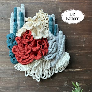 May include: A handmade crochet wall hanging in the shape of coral. The piece features various textures and colors, including teal, cream, red, and gray. The text "DIY Pattern" is in a white oval on the right side. The artwork is mounted on a wooden background.