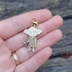 Może przedstawiać: Złoty charms przedstawiający postać z kreskówki z dużymi uszami, zielonymi oczami i niebieskim przedmiotem. Charms jest przymocowany do złotego zapięcia. Postać ma na sobie beżowy strój.