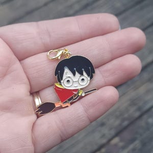 Puede incluir: Un dije dorado con un personaje de dibujos animados con cabello negro, gafas y una capa roja, montado en una escoba. El dije está sujeto a un cierre dorado. El dije se sostiene en una mano.