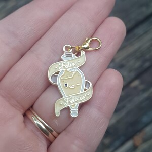 May include: A gold-coloured charm featuring a potion bottle design. The bottle is cream-coloured with a smiling face and the words "Felix Felicis" on a cream-coloured banner. The charm has a lobster clasp.