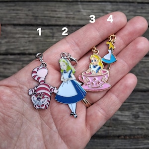Op de afbeelding: Vier metalen emaille bedels met personages uit Alice in Wonderland. Bedel 1 is een roze en wit gestreepte Cheshire Cat. Bedel 2 is Alice in een blauwe jurk. Bedel 3 is Alice in een roze theekopje. Bedel 4 is een silhouet van Alice in een blauwe jurk.