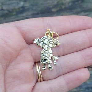 Op de afbeelding: Een goudkleurige bedel in de vorm van een gestileerd wezen met een geometrisch patroon op het lichaam. De bedel heeft een karabijnhaak voor bevestiging. De bedel wordt in een hand gehouden.