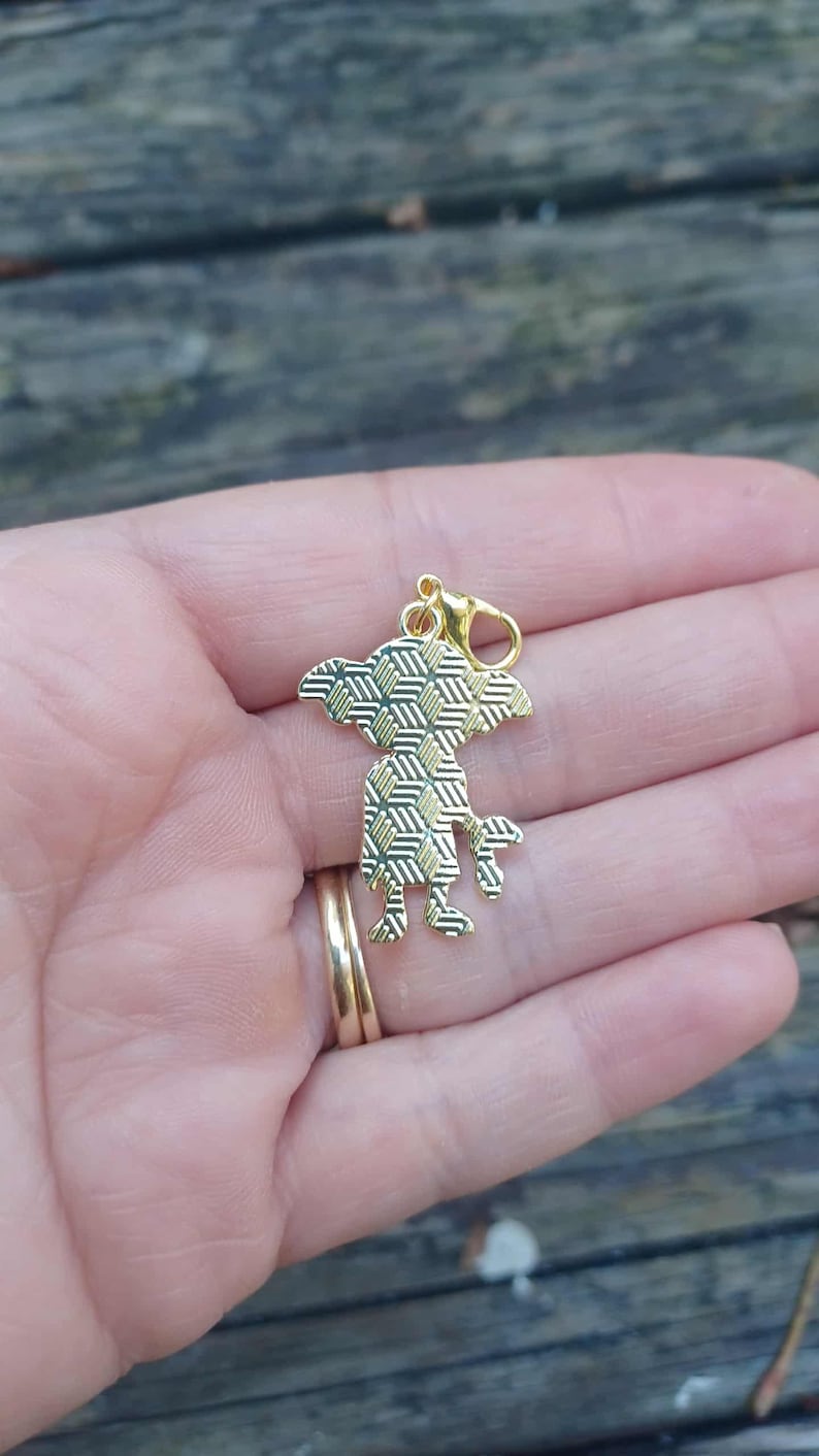 Op de afbeelding: Een goudkleurige bedel in de vorm van een gestileerd wezen met een geometrisch patroon op het lichaam. De bedel heeft een karabijnhaak voor bevestiging. De bedel wordt in een hand gehouden.