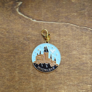 May include: A round charm with a gold-colored clasp featuring a Hogwarts castle design. The charm has a light blue background with the castle in brown and black, and the word "HOGWARTS" at the bottom.