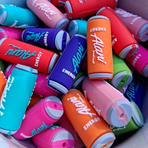 Op de afbeelding: Een verzameling kleine, cilindervormige siliconen bedels in de vorm van energiedrankblikjes in diverse kleuren zoals oranje, roze, blauw, paars en rood. Elke bedel is voorzien van de in reliëf aangebrachte witte tekst Alani Energy.