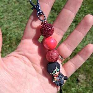 May include: A keychain featuring a black metal lobster clasp and a series of four beads. The beads include a dark red rhinestone sphere, a smooth red sphere, a glittery red sphere, and a character charm of a boy with black hair and glasses.