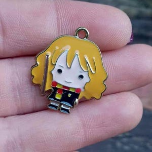 May include: A colourful enamel charm depicting a cartoon character with yellow hair, a red and black scarf, and a black outfit. The charm has a gold-coloured loop at the top for attaching to jewellery. The charm is held in a person's hand.