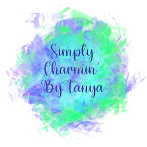 Op de afbeelding: Een aquarel-stijl ontwerp met een witte achtergrond. Het ontwerp toont een splash van groene, blauwe en paarse tinten. De tekst "Simply Charmin' By Tanya" is geschreven in een donkerblauwe, elegante scriptletter.