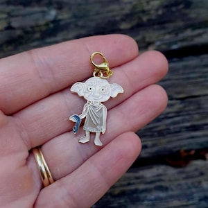 Op de afbeelding: Een goudkleurige bedel met een stripfiguur met grote ogen, puntige oren en een blauw voorwerp. De bedel is wit, lichtroze en goudkleurig. Hij is bevestigd aan een goudkleurige sluiting.
