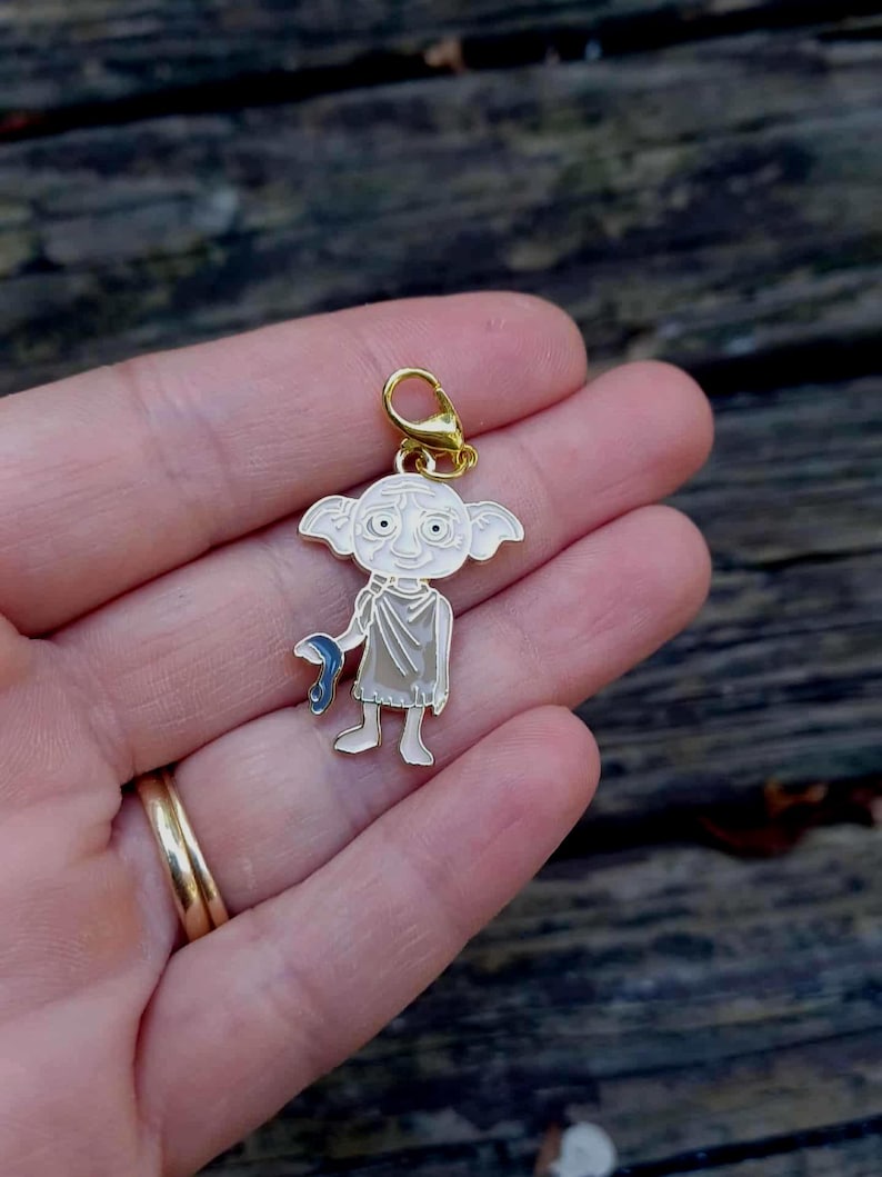 Op de afbeelding: Een goudkleurige bedel met een stripfiguur met grote ogen, puntige oren en een blauw voorwerp. De bedel is wit, lichtroze en goudkleurig. Hij is bevestigd aan een goudkleurige sluiting.