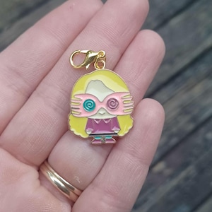Op de afbeelding: Een goudkleurige bedel met een stripfiguur met blond haar, een roze bril en een roze en paarse outfit. De bedel heeft een gouden sluiting en wordt in een hand gehouden.