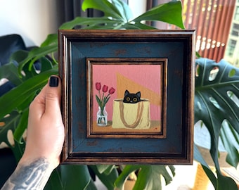 Puede incluir: Pintura enmarcada cuadrada que presenta un gato negro asomándose de una bolsa de mano amarilla. Un jarrón con tulipanes rojos a la izquierda. El fondo es rosa con un diseño geométrico. El marco es azul y marrón.