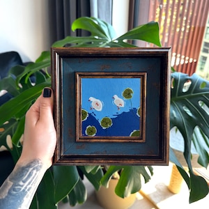 Op de afbeelding: Een vierkant schilderij in een donkerbruine en blauwe verweerde lijst met twee witte eenden die zwemmen op een felblauwe vijver met donkerblauwe waterrimpels en verschillende groene waterleliebladeren verspreid over het wateroppervlak.