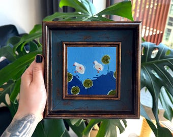 Puede incluir: Una pintura cuadrada en un marco desgastado de color azul y marrón oscuro que muestra dos patos blancos nadando en un estanque azul brillante con ondas de agua azul oscuro y varios nenúfares verdes esparcidos por la superficie.